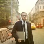Confident well dressed male entrepreneur in elegant suit and eyeglasses standing with netbook near car on road and looking at camera